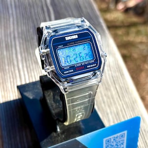 K&ouml;nnte beinhalten: Transparente Digitaluhr mit blauem Zifferblatt, das die Uhrzeit 10:26:26 anzeigt. Die Uhr hat ein transparentes Armband und eine schwarze L&uuml;nette. Das Zifferblatt tr&auml;gt die Aufschrift "SKMEI", "ALARM CHRONOGRAPH", "WATER 50M RESIST".