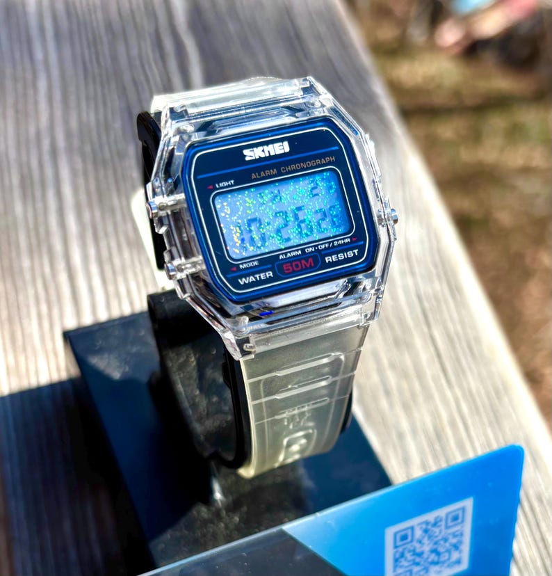K&ouml;nnte beinhalten: Transparente Digitaluhr mit blauem Zifferblatt, das die Uhrzeit 10:26:26 anzeigt. Die Uhr hat ein transparentes Armband und eine schwarze L&uuml;nette. Das Zifferblatt tr&auml;gt die Aufschrift "SKMEI", "ALARM CHRONOGRAPH", "WATER 50M RESIST".
