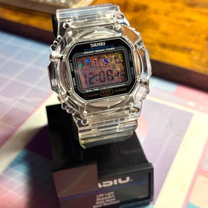 K&ouml;nnte beinhalten: Eine transparente Digitaluhr mit schwarzem Zifferblatt, das die Uhrzeit 12:08:4 anzeigt. Die Uhr hat ein quadratisches Zifferblatt und ein transparentes Armband. Die Uhr steht auf einem schwarzen St&auml;nder. Auf dem Zifferblatt steht "SKMEI" und "COUNT DOWN TIMER".