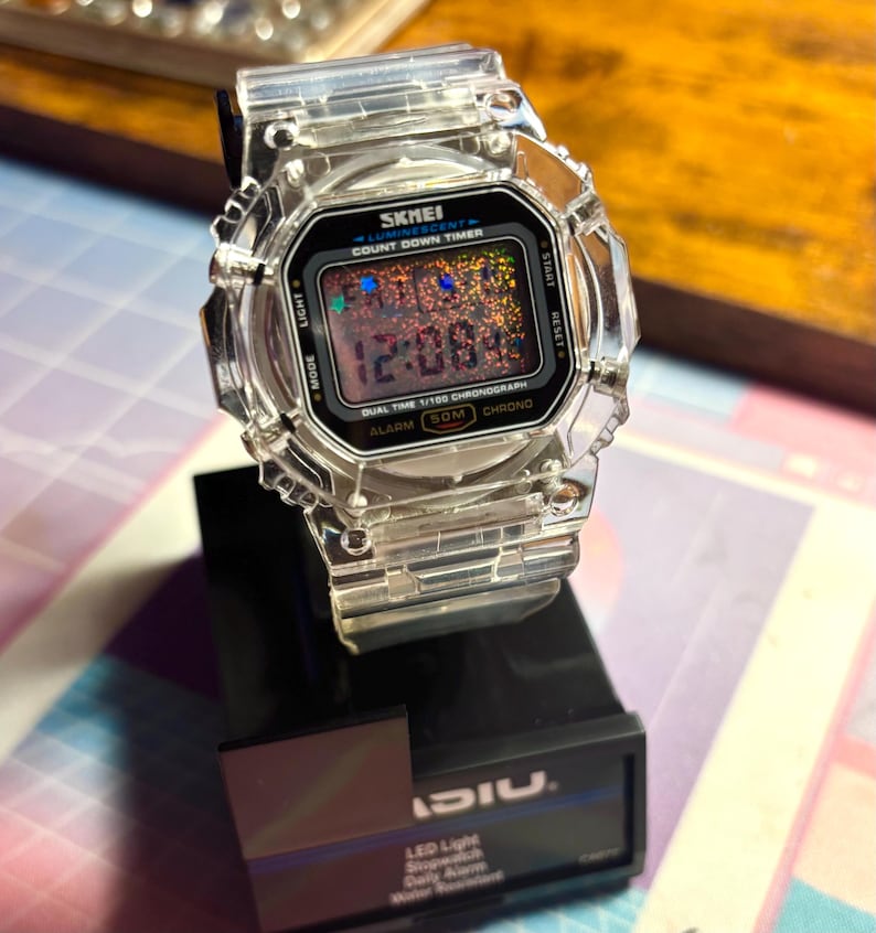 K&ouml;nnte beinhalten: Eine transparente Digitaluhr mit schwarzem Zifferblatt, das die Uhrzeit 12:08:4 anzeigt. Die Uhr hat ein quadratisches Zifferblatt und ein transparentes Armband. Die Uhr steht auf einem schwarzen St&auml;nder. Auf dem Zifferblatt steht "SKMEI" und "COUNT DOWN TIMER".