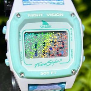 Puede incluir: Un reloj digital con caja blanca y azul claro que presenta una pantalla holográfica. La esfera dice NIGHT VISION, SHARK, FreeStyle y 300 FT. La correa es azul claro con un logotipo de aleta de tiburón blanco y el texto SHARK CLIP.