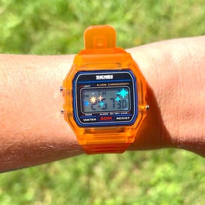 May include: A bright orange digital watch with a translucent resin case and strap. The rectangular face features the brand name SKMEI, the text ALARM CHRONOGRAPH, and WATER 50M RESIST. The digital display shows the time 2:57 and date 30.