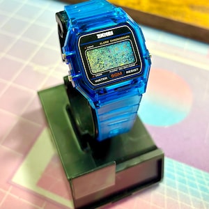 Puede incluir: Un reloj digital SKMEI azul transparente con una esfera negra, exhibido en un soporte negro. El reloj presenta una pantalla digital con el texto "SKMEI ALARM CHRONOGRAPH" y "WATER 50M RESIST". La correa y la caja del reloj son de un azul vibrante.