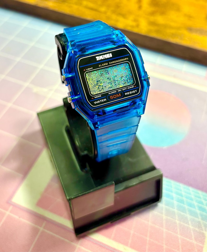 Puede incluir: Un reloj digital SKMEI azul transparente con una esfera negra, exhibido en un soporte negro. El reloj presenta una pantalla digital con el texto "SKMEI ALARM CHRONOGRAPH" y "WATER 50M RESIST". La correa y la caja del reloj son de un azul vibrante.