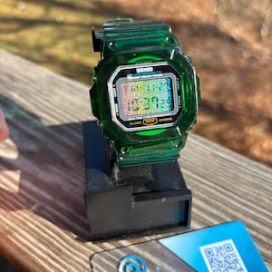 May include: A transparent green digital watch with a black display, resting on a black stand. The watch face displays the time and date. The watch is labeled "SKMEI" and has several function buttons. A blue and white card with a QR code and the VaporFine logo is next to the watch.