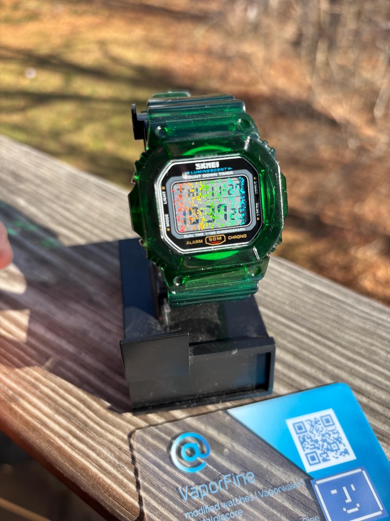 May include: A transparent green digital watch with a black display, resting on a black stand. The watch face displays the time and date. The watch is labeled "SKMEI" and has several function buttons. A blue and white card with a QR code and the VaporFine logo is next to the watch.