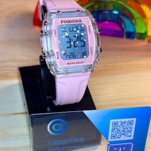 May include: A pink digital watch with a clear rectangular case and a pink strap. The watch face displays the time, date, and the word "PINDOWS". The watch is water resistant and has multiple buttons. The watch is displayed on a black stand.