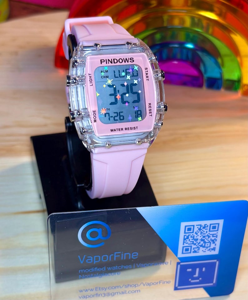 May include: A pink digital watch with a clear rectangular case and a pink strap. The watch face displays the time, date, and the word "PINDOWS". The watch is water resistant and has multiple buttons. The watch is displayed on a black stand.