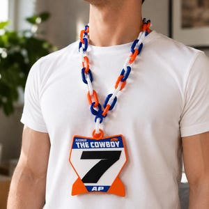 May include: A necklace featuring a thick chain of interlocking orange, white, and blue plastic links. A large, shield-shaped pendant hangs from the chain, displaying the text PLESSINGER, THE COWBOY, and AP, with a prominent black number 7.
