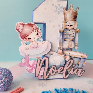 May include: A birthday-themed decoration featuring a ballerina, a nutcracker, and the name "Noelia". The number "1" is present, along with snowflakes and a blue "Happy Birthday" headband. The color palette includes pink, blue, and white.