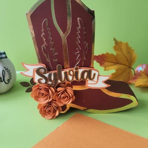 May include: A three-dimensional paper craft in the shape of a cowboy boot, colored in deep maroon with gold metallic trim. The boot features a banner across the front with the name Sylvia in dark brown script, accented by three paper roses.
