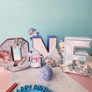 May include: A festive birthday decoration featuring the word "ONE" in large, decorative letters. The letters are adorned with winter-themed cutouts, including a snowman, Santa, and a ballerina. A "Happy Birthday" headband and a glitter ball complete the scene.