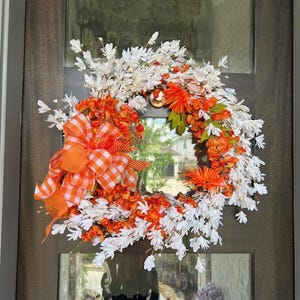 Puede incluir: Una corona decorativa con elementos florales blancos, naranjas y verdes. La corona presenta un gran lazo a cuadros naranja y blanco. La corona se exhibe en una puerta marrón con paneles de vidrio.