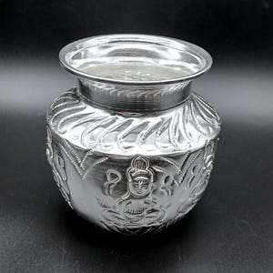 May include: A silver-colored, ornate pot with a wide rim and detailed relief carvings. The pot features a rounded body with floral and figure designs, and a smooth, reflective surface. The pot is likely made of metal.