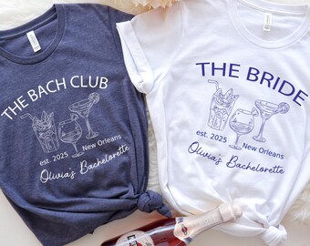 May include: Two t-shirts with a bachelorette party theme. The blue shirt says "The Bach Club" and the white shirt says "The Bride". Both shirts feature a design of three cocktails and the text "est. 2025 New Orleans Olivia's Bachelorette."