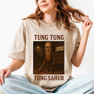 May include: Beige t-shirt featuring a graphic of a cartoon character holding a bat, with the text "TUNG TUNG" above and "TUNG SAHUR" below. The graphic is a sepia tone, and the shirt is a neutral color.