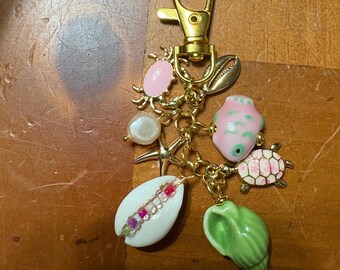 Könnte beinhalten: Ein goldfarbener Schlüsselanhänger mit Karabinerverschluss, verziert mit verschiedenen Anhängern. Zu den Anhängern gehören ein rosa Seestern, eine weiße Muschel mit bunten Perlen, eine grüne Muschel, eine rosa Krabbe, ein rosa Fisch, eine Perle und eine rosa-weiße Schildkröte.