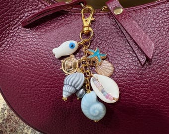 Könnte beinhalten: Ein goldfarbener Schlüsselanhänger mit Anhängern zum Thema Meer. Die Anhänger umfassen einen weißen Fisch mit blauem Auge, einen blauen Seestern und Muschelanhänger in Weiß, Blau und Grau. Der Anhänger ist an einer bordeauxfarbenen Ledertasche befestigt.
