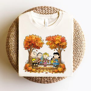 May include: A cream-colored t-shirt featuring a graphic print of the Peanuts characters sitting on a wooden bench under autumn trees. The design includes Charlie Brown, Linus, Sally, Snoopy, Woodstock, and Lucy holding mugs amidst orange pumpkins.