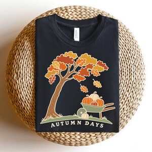May include: A black t-shirt featuring a graphic of a tree with orange, yellow, and red leaves, a wheelbarrow filled with pumpkins, and the text "AUTUMN DAYS" printed in white at the bottom. The design includes falling leaves and grass.