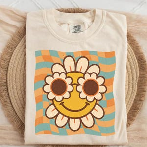May include: Cream-colored t-shirt featuring a retro-style smiley face with flower-shaped sunglasses. The graphic is set against a wavy, checkered background in shades of orange and teal. The shirt has a classic crew neck and a Comfort Colors tag.