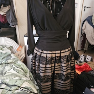 May include: A black dress with a V-neck bodice and long sleeves. The waist is cinched with a wide band, and the skirt features alternating stripes of beige fabric and black lace. The dress is displayed on a dress form.