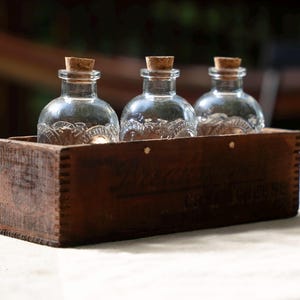 May include: Three clear glass bottles with cork stoppers sit in a rustic, dark brown wooden box. The bottles have a decorative pattern on the lower half. The box has a rectangular shape and is placed on a white surface.