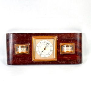 Könnte beinhalten: Vintage Wetterstation aus Holz mit zentralem Barometer und zwei kleineren Messgeräten. Das Barometer hat ein weißes Zifferblatt mit schwarzen Ziffern und einen goldfarbenen Rahmen. Das Holz hat eine dunkle, rotbraune Oberfläche.