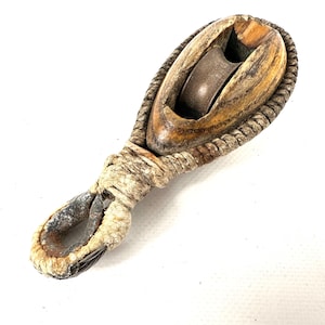 May include: A vintage wooden pulley with a metal wheel and rope detailing. The pulley is made of weathered wood with a natural finish, and the rope is a light beige colour. The metal wheel shows signs of age and wear.