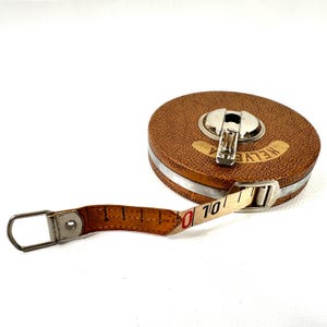 May include: A vintage brown leather Helvetia measuring tape with a metal handle and a white and tan measuring tape extending out. The tape reads "10" at the end. The tape measure is on a white background.