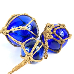 Può includere: Tre galleggianti da pesca in vetro blu cobalto di varie dimensioni, ciascuno racchiuso in una rete di corda annodata di colore naturale. I galleggianti sono disposti su uno sfondo bianco, mettendo in risalto il loro colore e la loro consistenza vibranti.