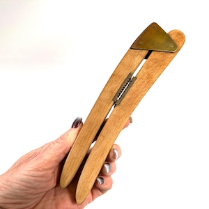 Può includere: Un tendiabiti in legno con un triangolo in ottone e una cerniera in metallo. Il tendiabiti è realizzato in legno chiaro ed è progettato per allungare i vestiti. Lo strumento è tenuto in mano su sfondo bianco.