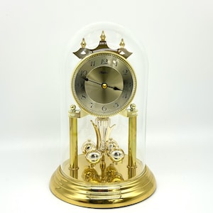 Può includere: Un orologio da mensola dorato con una cupola di vetro. Il quadrante dell'orologio è dorato con numeri e lancette nere. La base e gli elementi decorativi sono dorati. L'orologio ha un pendolo con tre sfere argentate.