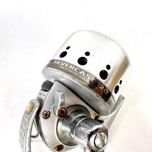 May include: Silver fishing reel with the text "SILVERCAST 120" and "12lb.(0.35mm)-100yds.(90m)". The reel is silver with black circular holes on the top. The reel has a handle and other mechanical parts.