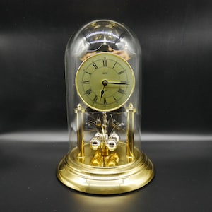 Può includere: Un orologio dorato con una cupola di vetro. Il quadrante è verde chiaro con numeri romani e lancette nere. Un pendolo con tre sfere argentate è appeso sotto. L'orologio poggia su una base dorata.