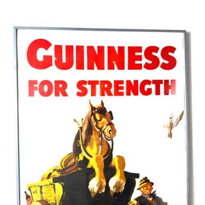 May include: Framed vintage Guinness advertisement. The red text reads "GUINNESS FOR STRENGTH" above a horse-drawn cart illustration. The horse is brown and gold, and a man in a hat is visible below the cart.