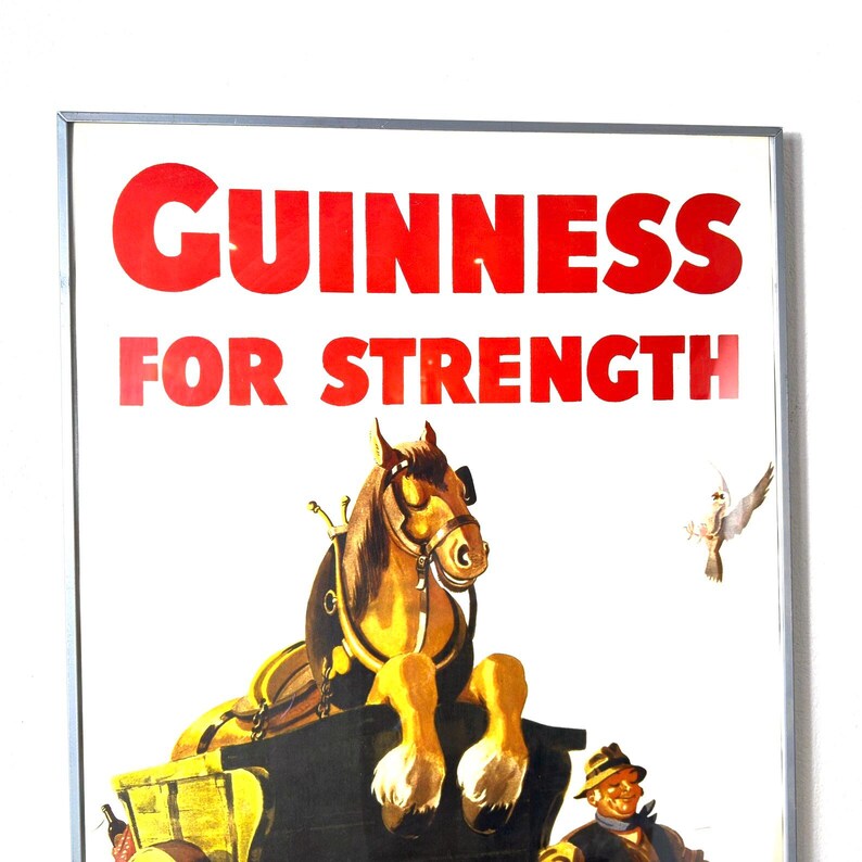 May include: Framed vintage Guinness advertisement. The red text reads "GUINNESS FOR STRENGTH" above a horse-drawn cart illustration. The horse is brown and gold, and a man in a hat is visible below the cart.