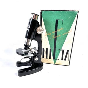 Puede incluir: Un microscopio negro vintage con detalles plateados y una caja a juego. La caja presenta un diseño de triángulo verde con las palabras "Today investigator Tomorrow scientist" y la marca "MILTA".