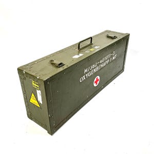May include: An olive green rectangular metal box with a black handle and metal latches. The box has white text and a red cross symbol. A yellow and black warning label is on the side. The box is likely for medical supplies.
