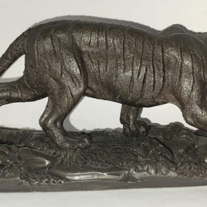 Op de afbeelding: Een bronzen beeld van een tijger die op een getextureerde basis loopt. De tijger is afgebeeld in een realistische stijl met gedetailleerd haar en strepen.