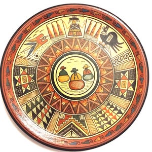 May include: A circular, hand-painted ceramic plate with a reddish-brown rim and detailed designs. The plate features a central circle with three figures, surrounded by geometric patterns in yellow, orange, brown, and black. The plate has a traditional, cultural theme.