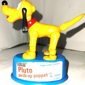May include: A yellow Pluto push-up puppet toy with black ears, a red collar, and a blue base. The toy is labeled "Gabriel" and "Pluto push-up puppet". The base also includes the text "Ages over 3 years."