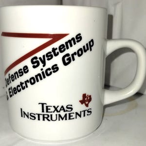 May include: White ceramic mug with a curved handle. The mug features the text "Defense Systems & Electronics Group" and "Texas Instruments" in black, with a red angled design. A red logo is also present.