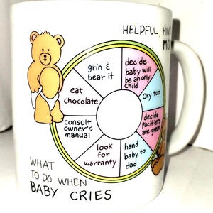 May include: White ceramic mug with a cartoon bear illustration and text. The mug features a circular diagram with advice on what to do when a baby cries, including "eat chocolate" and "hand baby to dad."