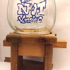 Op de afbeelding: Een heldere glazen pot met de tekst "The NUT Vendor" in blauwe letters staat op een houten standaard. De standaard is gemaakt van onbewerkt hout en heeft een kleine lade. De pot heeft een afgeronde vorm.