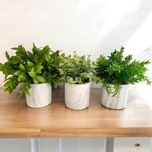 May include: Three potted artificial plants with varying shades of green foliage. The plants are in white ceramic pots with a marble-like pattern. The pots are arranged on a light brown wooden surface.