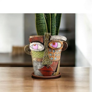 May include: A decorative ceramic face planter with a unique, abstract design. The planter features a face with large eyes, a nose, and lips, with a variety of colors including green, red, and brown. A green plant is inside the planter.