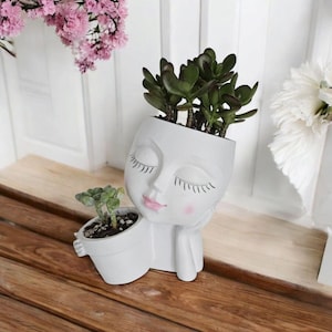 May include: A white, face-shaped planter with closed eyes, pink lips, and blush. The planter holds a green succulent. A smaller planter with a succulent is held in the planter's hand. The planter is on a wooden surface.
