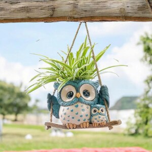 May include: A hanging owl planter with a large owl and a smaller owl, both in shades of blue and white, are perched on a wooden swing. The planter holds a green plant with long, slender leaves. The swing is suspended by rope.