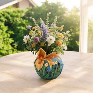May include: A colorful floral arrangement in a decorative vase. The vase is blue with orange and green accents, featuring an iris design. The flowers include white daisies, purple, yellow, and orange blooms. The arrangement sits on a light-colored wooden table.